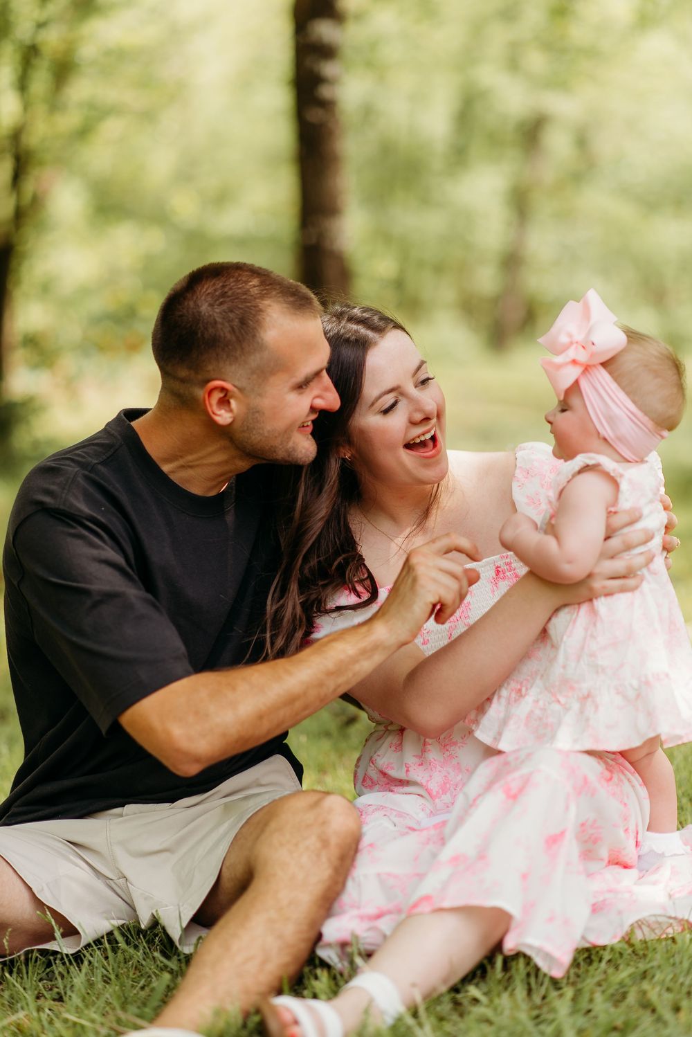 A loving family moment captured outdoors on the grass with parents and baby wearing pink dresses in a natural setting.