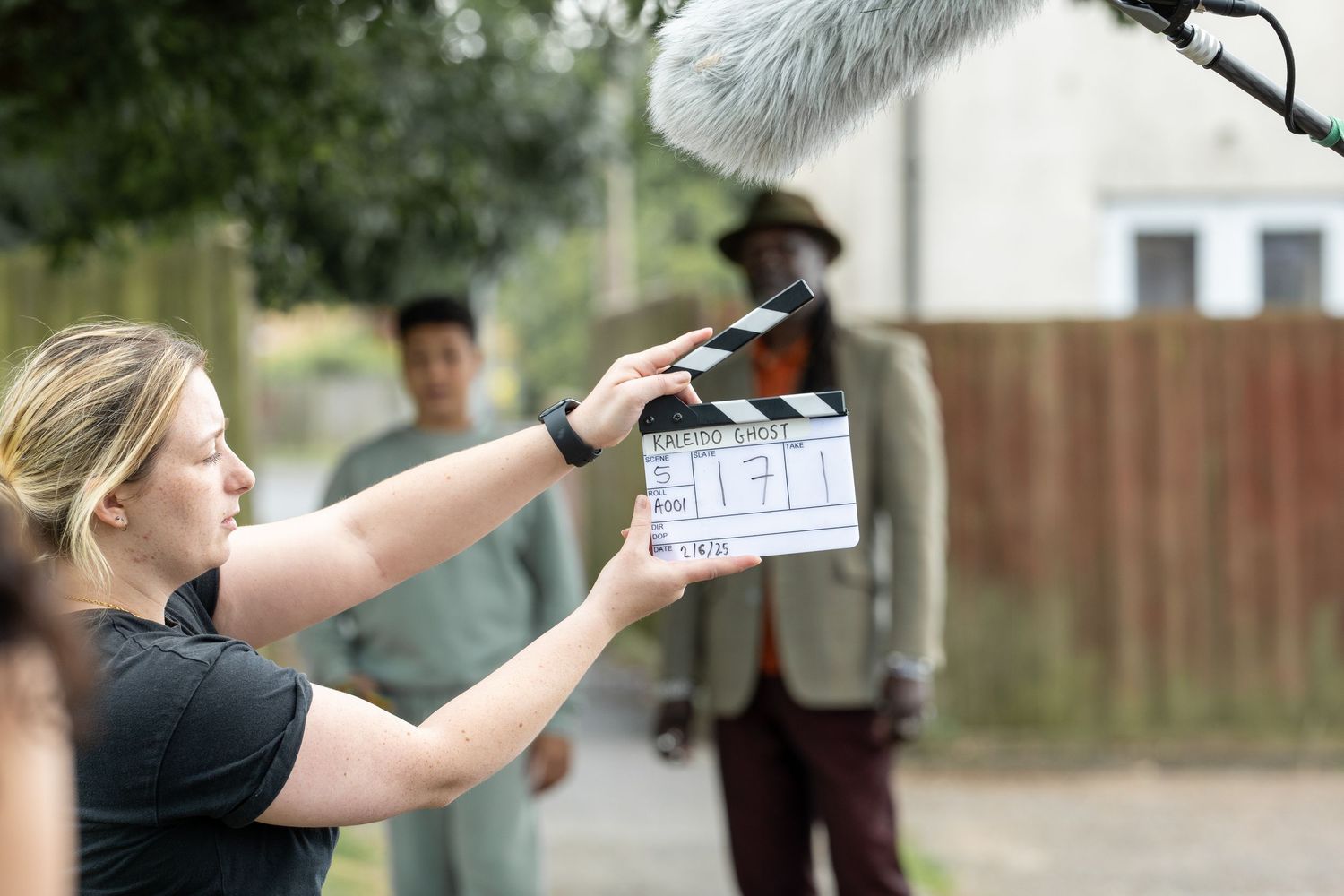 Film crew member holding a movie slate clapperboard during outdoor production shoot with equipment visible in background.