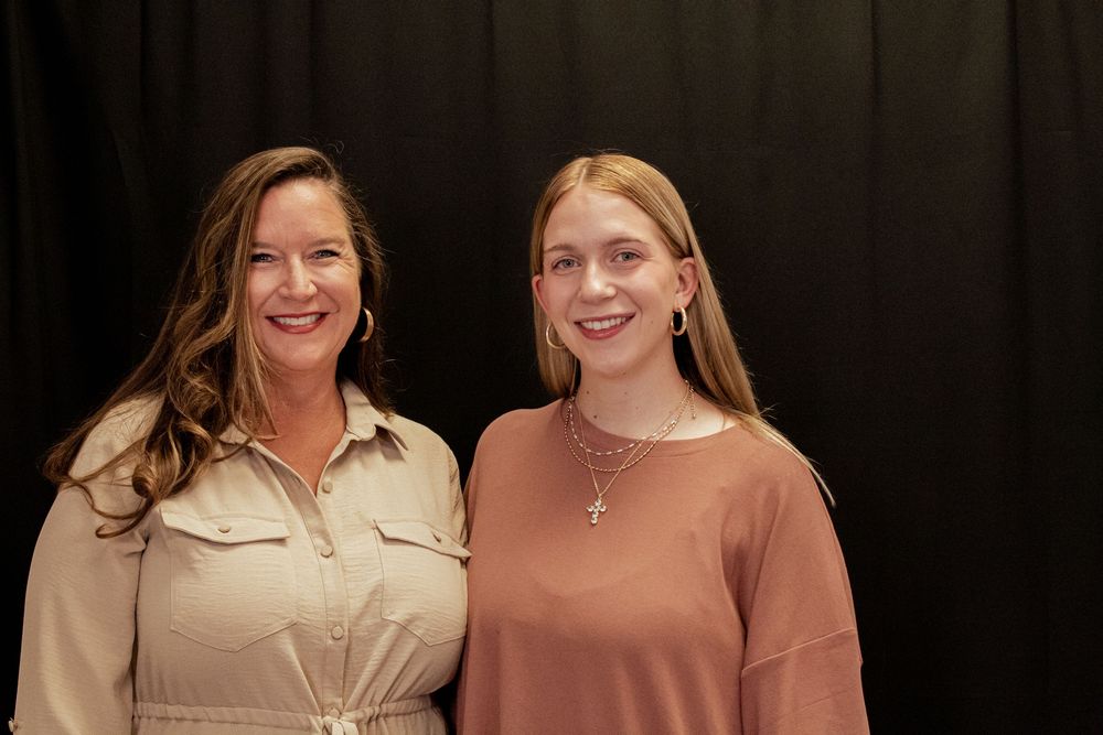 Two people stand together smiling warmly against a dark background.