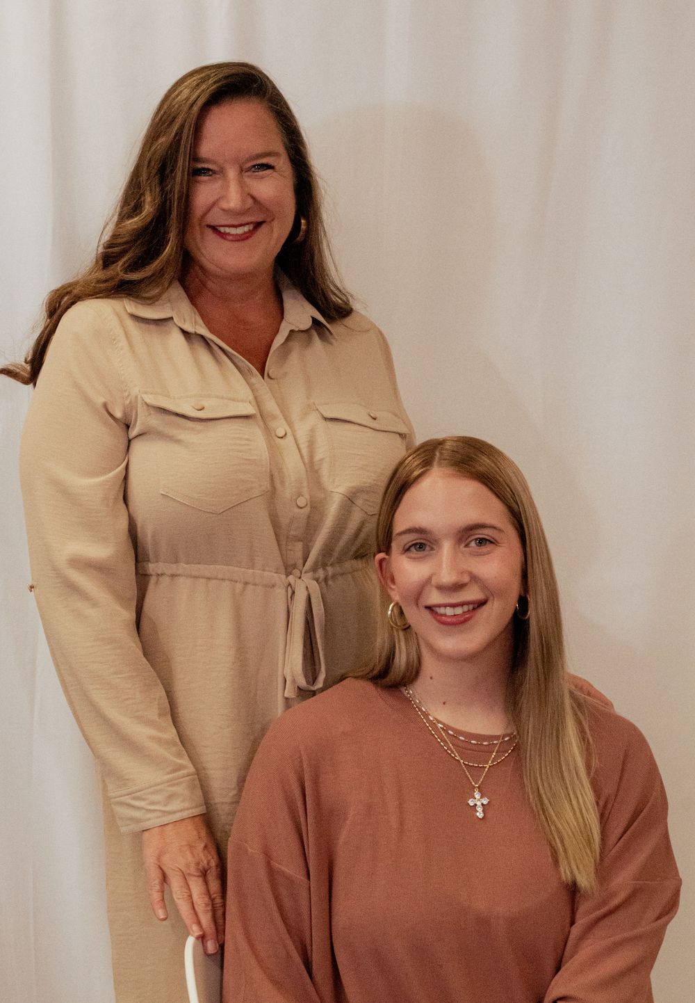 A pair wearing coordinating beige and brown outfits pose together while smiling warmly for the camera.
