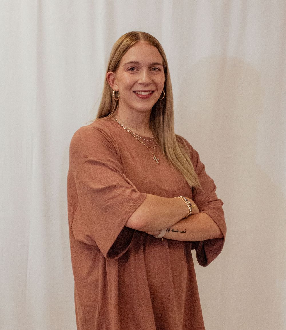 Person wearing a brown long-sleeved dress stands with arms crossed against white curtain background.