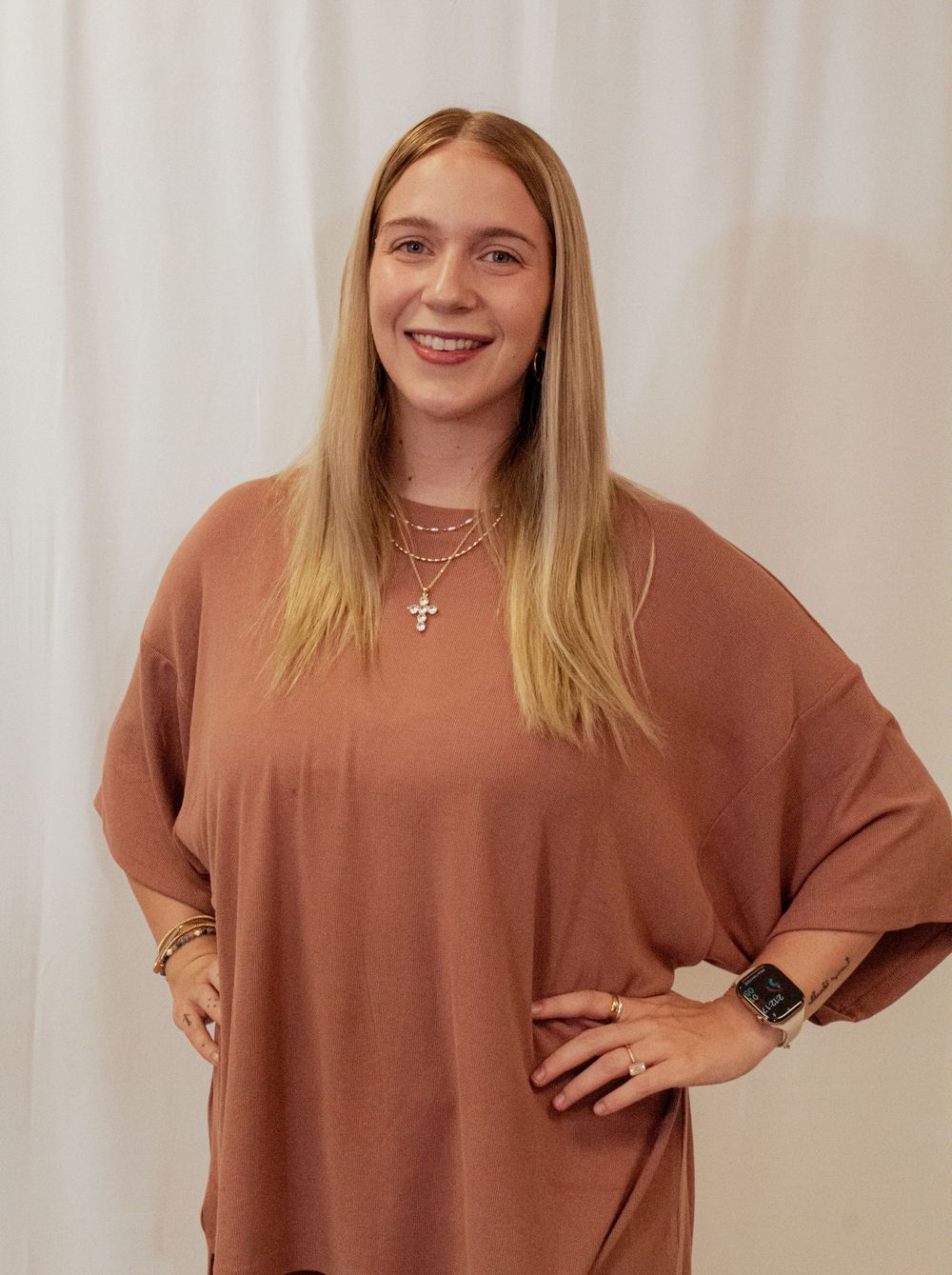 Person wearing a casual brown top with long blonde hair and delicate cross necklace against white background.