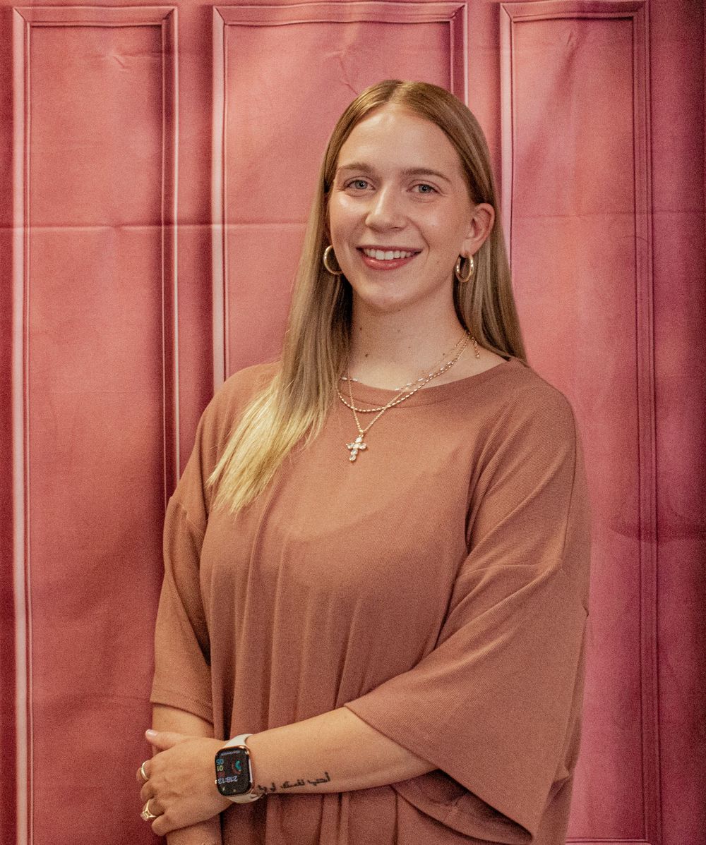 Portrait against pink wall wearing brown long sleeve top, cross necklace, hoop earrings and smart watch.