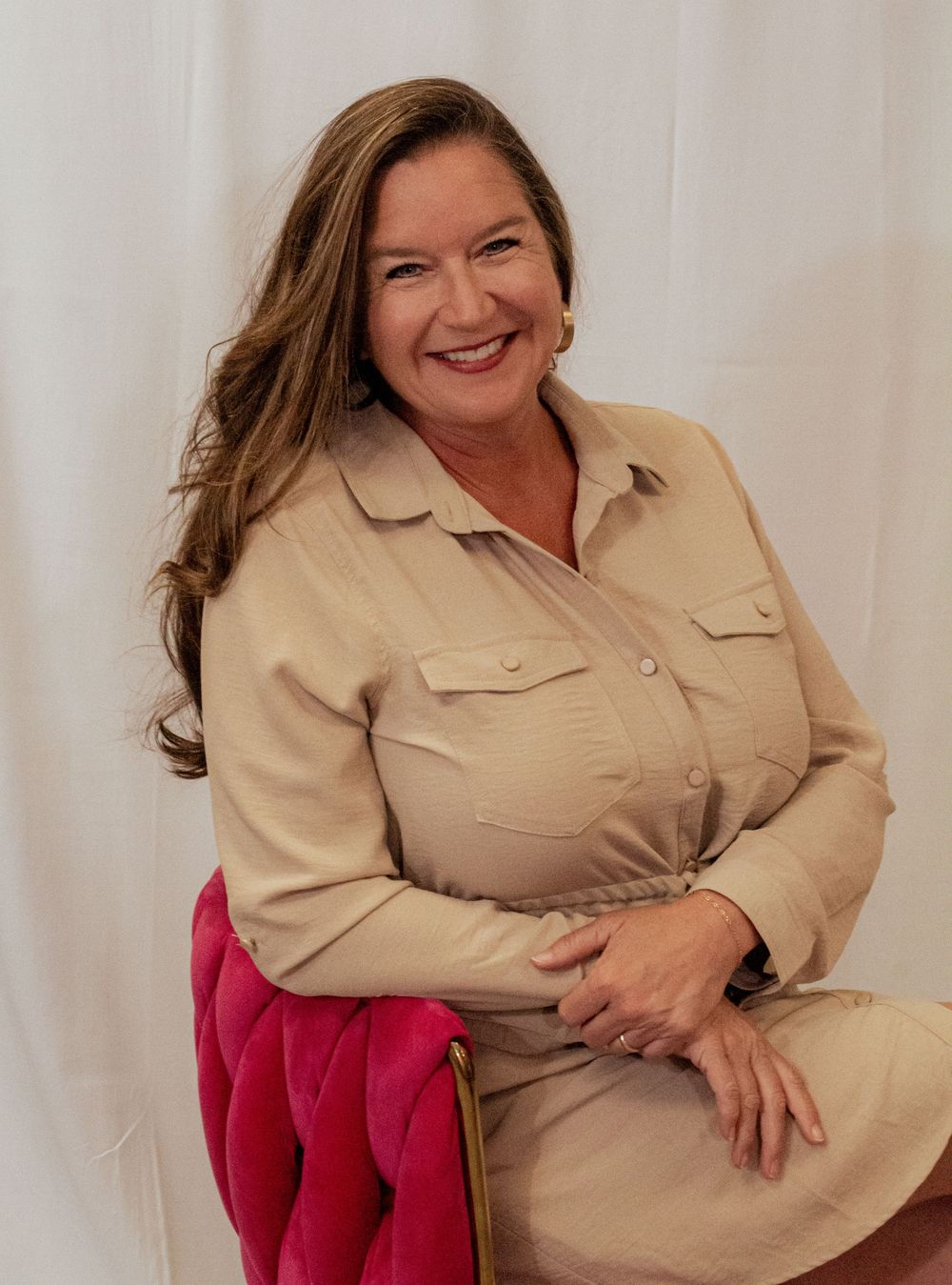 A person wearing a beige button-up shirt and red blazer poses against a white backdrop for a professional headshot.
