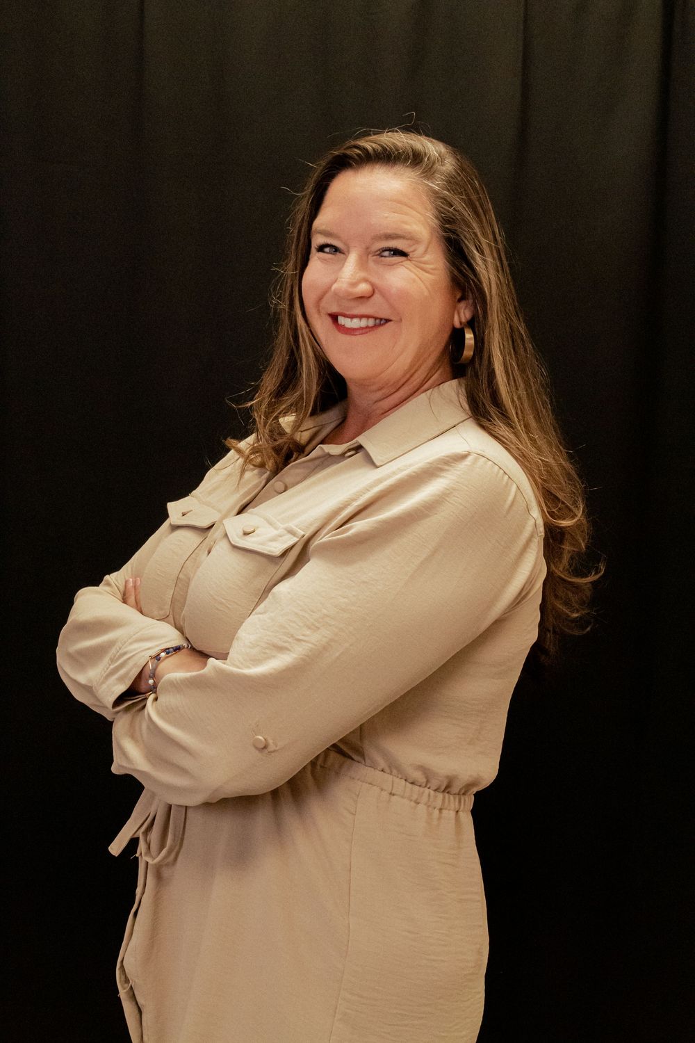 Portrait of a person in a beige dress smiling confidently with arms crossed against a dark background.