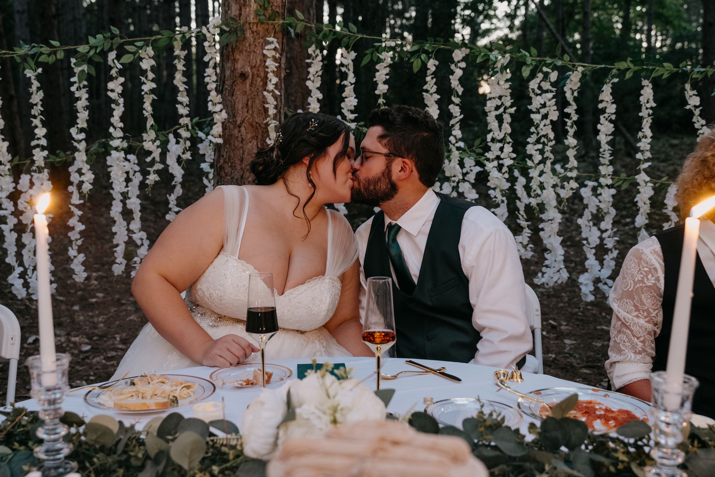 Couple sharing a romantic moment during an outdoor dinner under string lights and hanging greenery decorations.