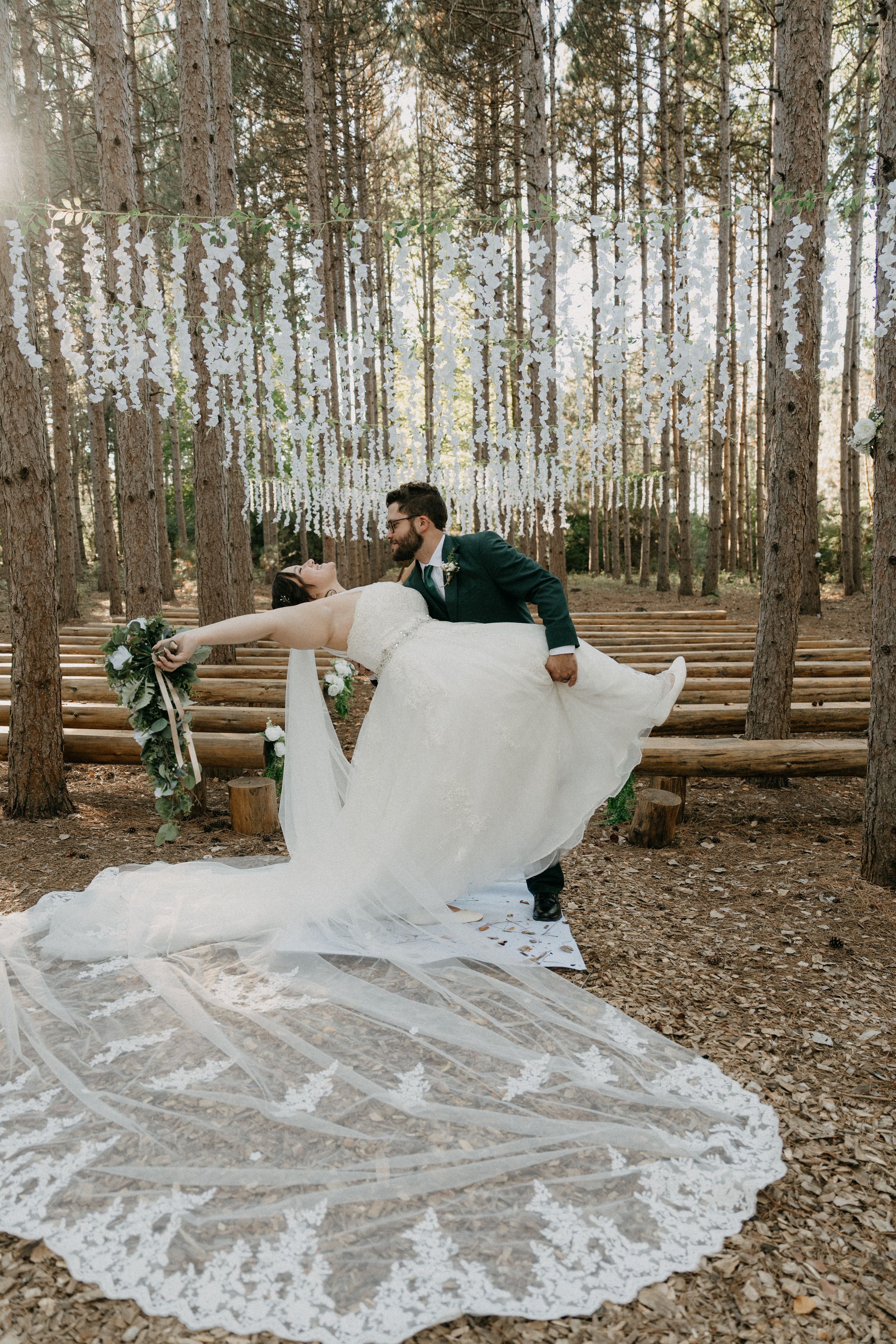 A wedding dress with a long lace train spreads across the forest floor between tall pine trees in a rustic woodland setting.