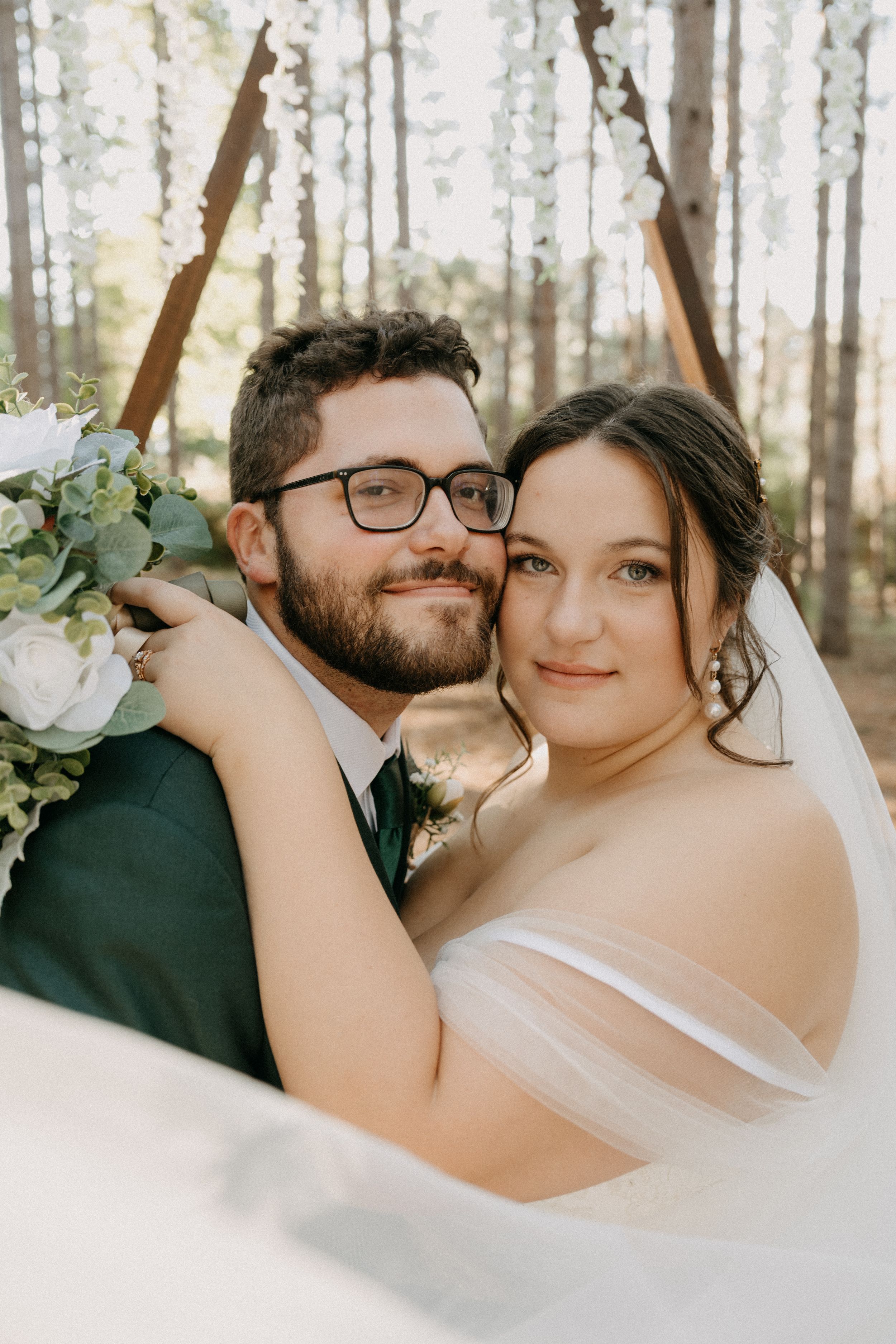 A romantic wedding portrait of a couple embracing in a sunlit forest with white floral bouquet details.