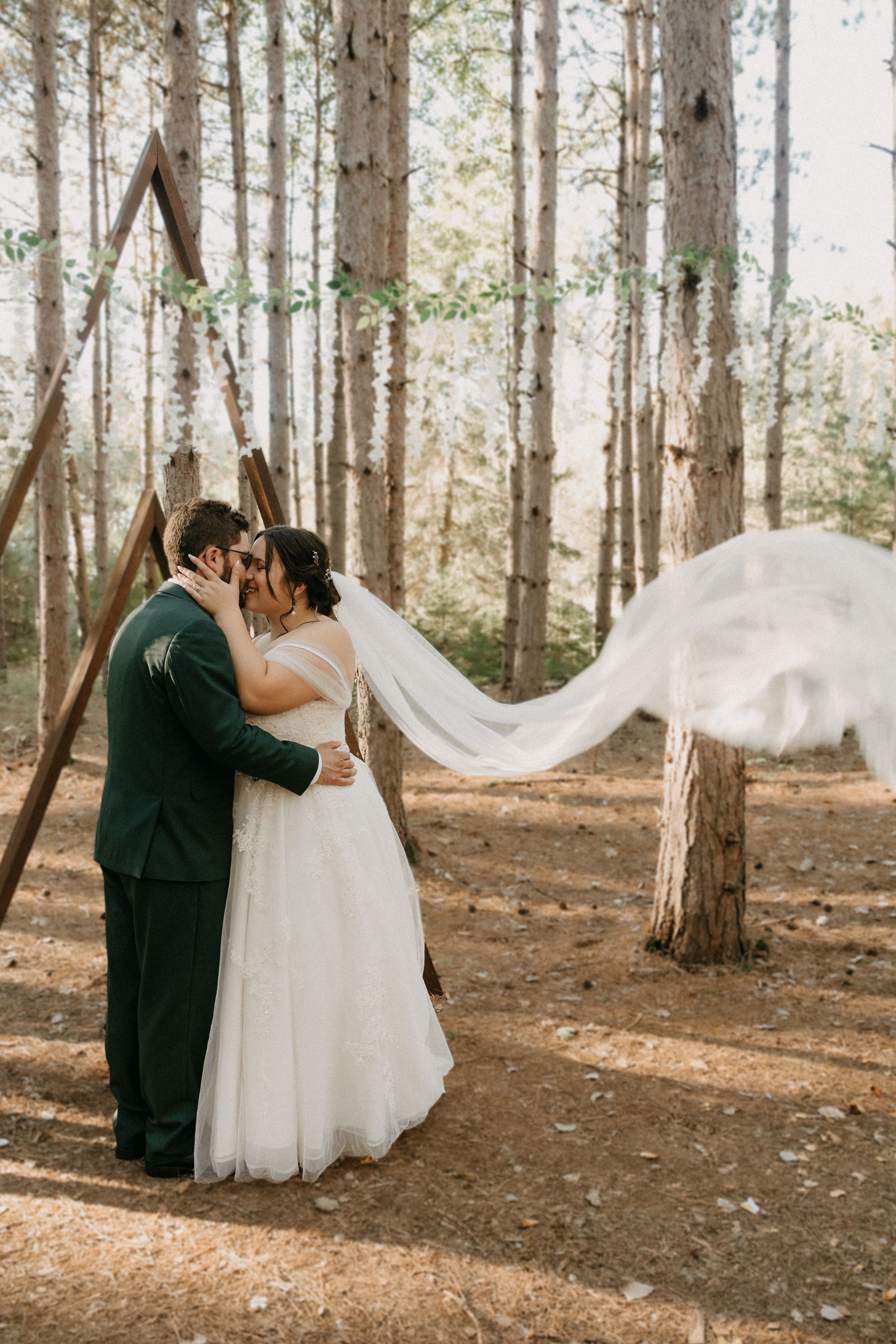 Romantic couple in formal wedding attire embrace among tall pine trees in a sunlit forest setting.