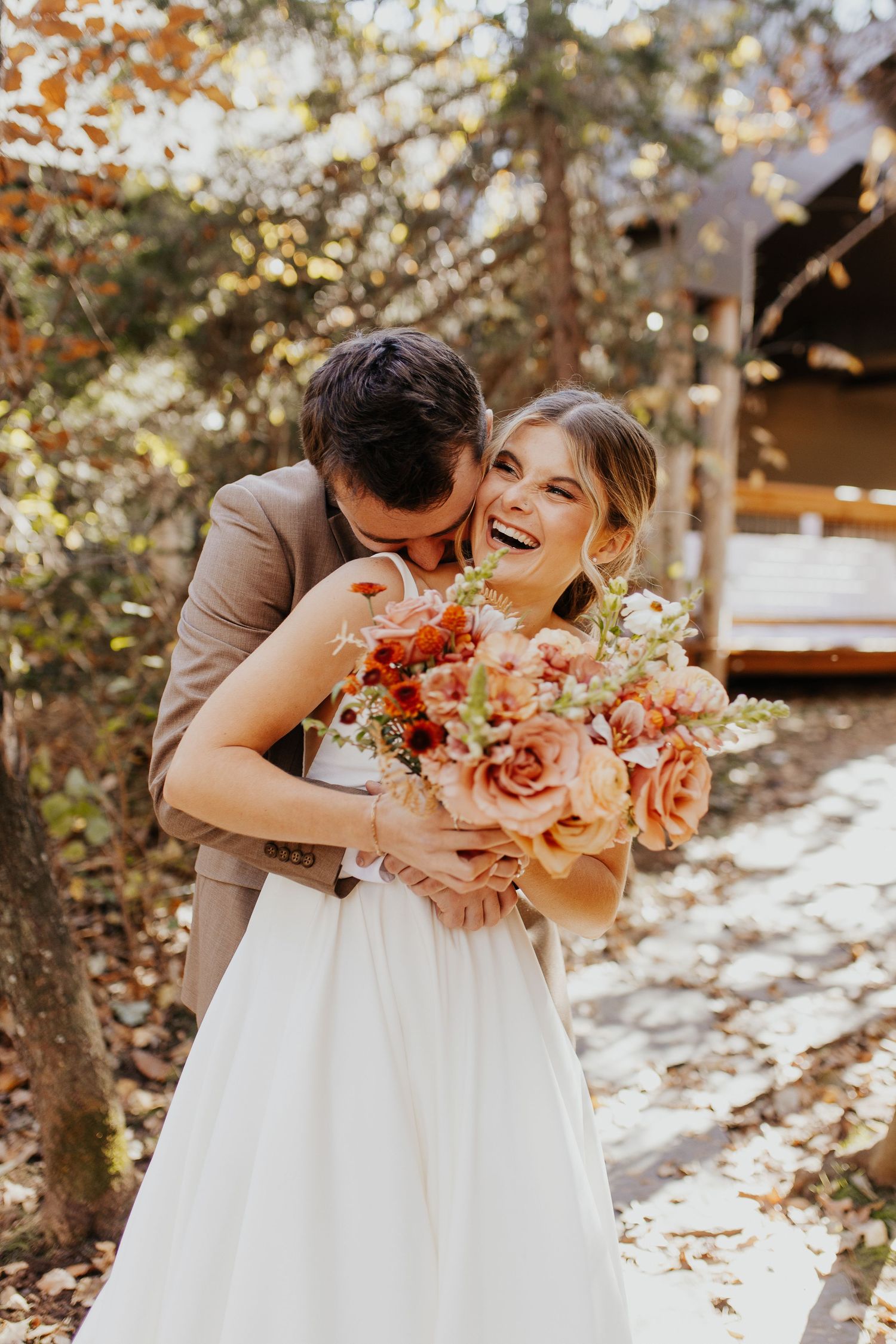 A couple embraces and laughs together in a romantic garden setting with coral and peach colored flowers.