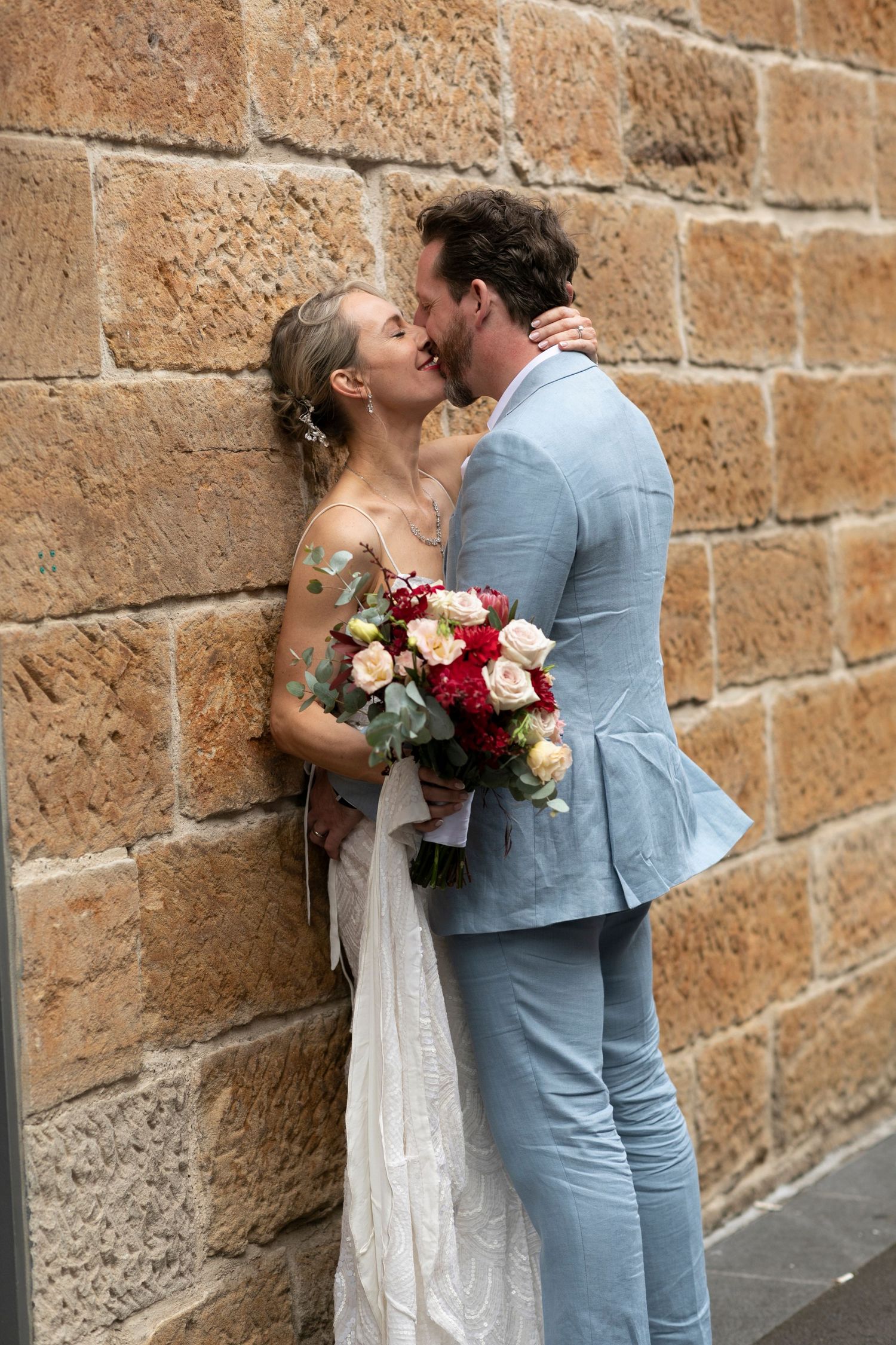 A bride in a white dress holds a colorful bouquet while kissing against a rustic sandstone wall.
