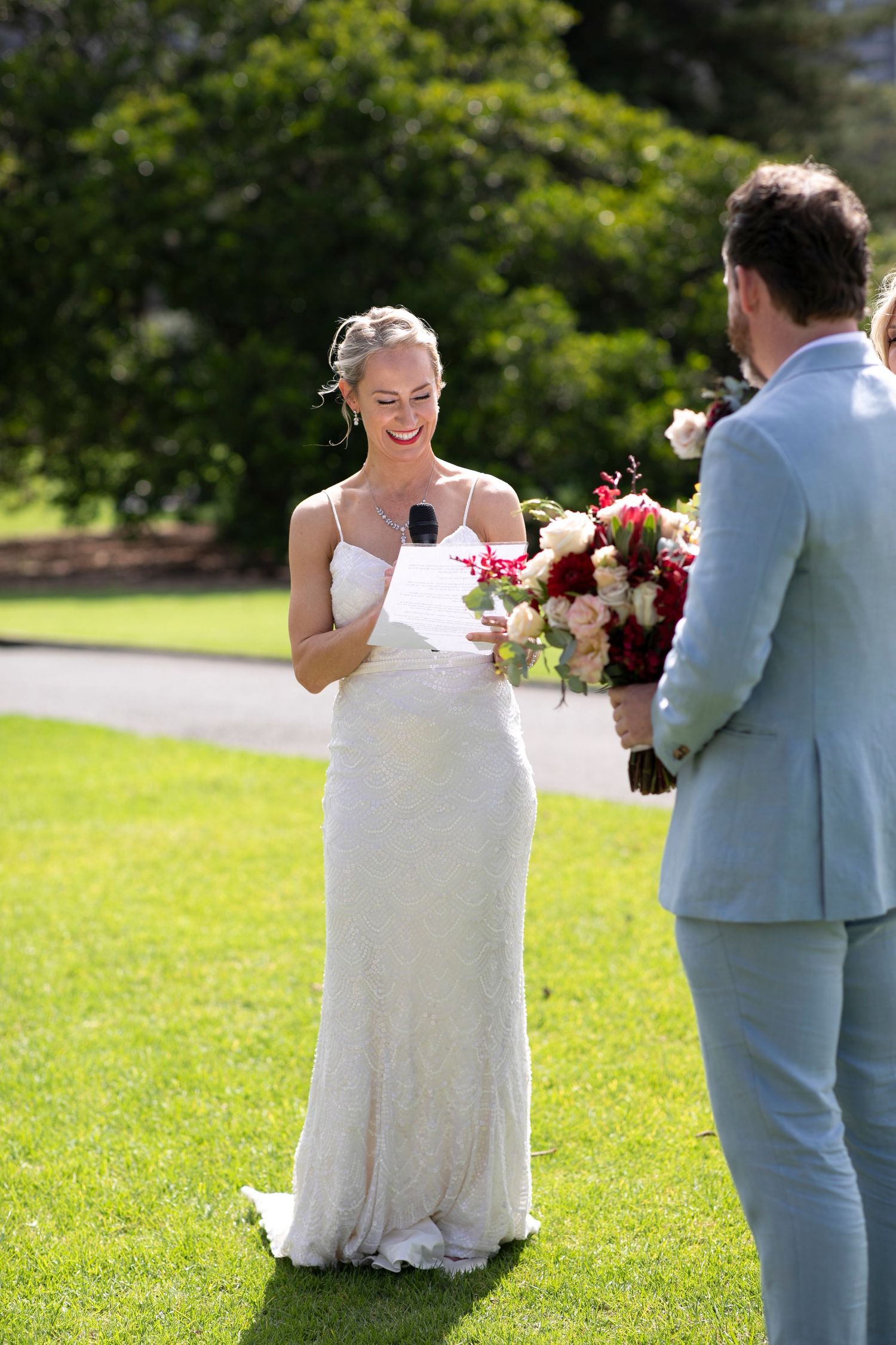 A couple exchanging vows outdoors on a sunny day with a lush green lawn and trees in the background.