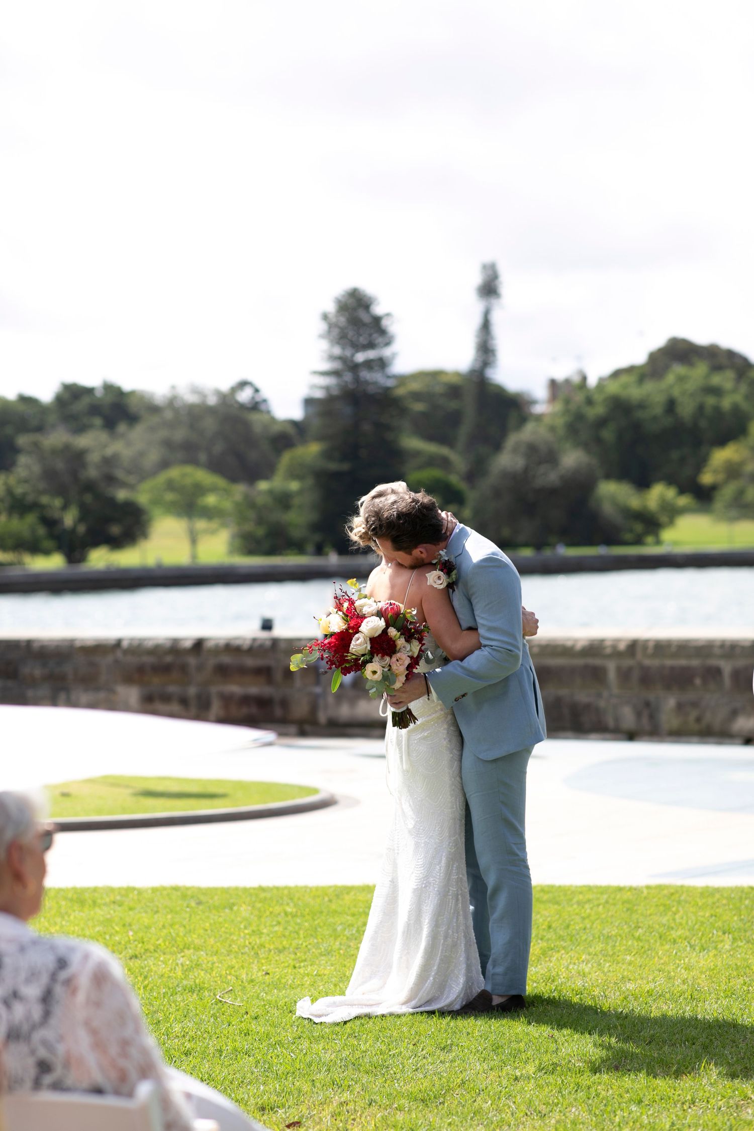 Couple share an intimate moment in formal attire on a manicured lawn with water and trees in background.