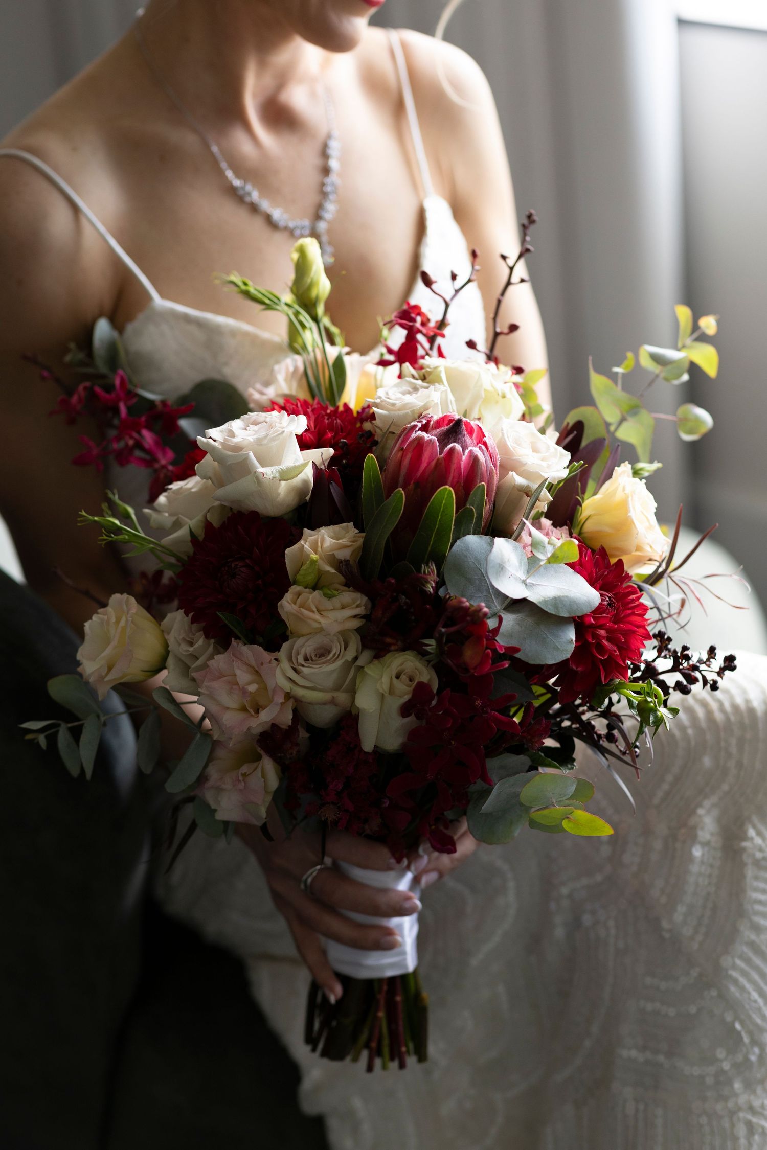 Dark and moody bridal bouquet featuring burgundy and cream roses with greenery in dramatic lighting.