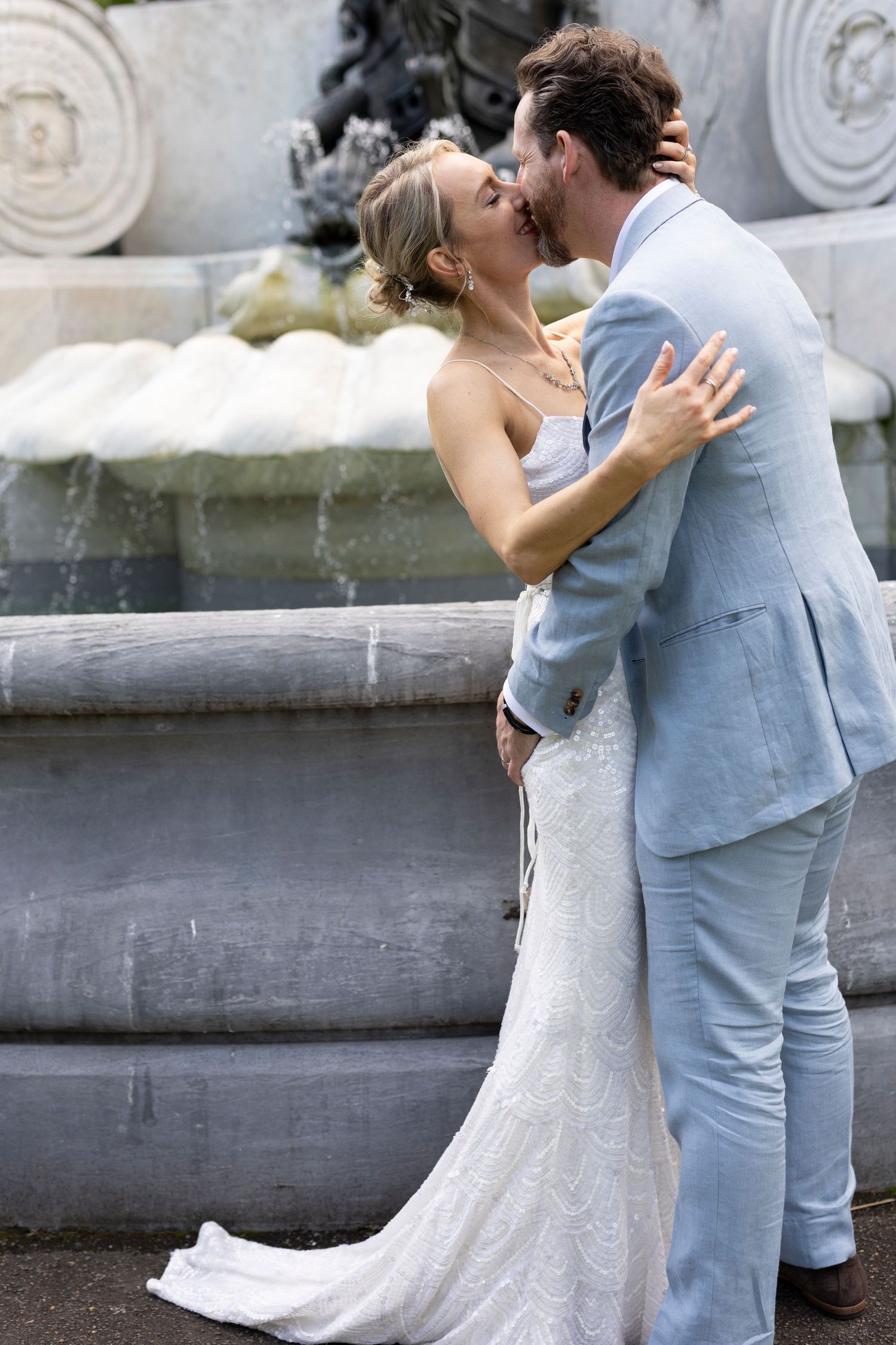 A couple in wedding attire share an intimate kiss in front of a stone fountain, with the bride in white lace and groom in blue suit.