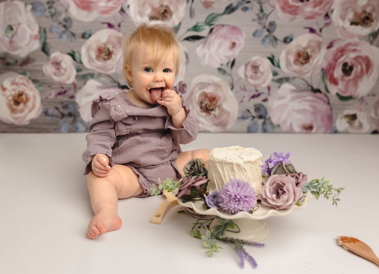 Somerset Portraits captures playful cake smash moment with one-year-old girl in purple outfit and floral backdrop in Charlottesville studio.