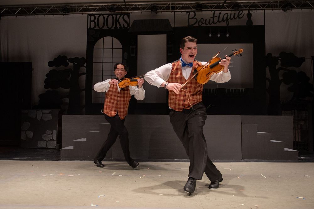 Two performers in matching plaid vests dance energetically in front of a bookstore storefront during a theatrical show.