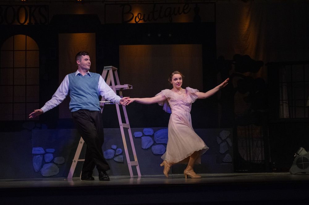 Two performers dance together on a theatrical stage with a ladder prop and dramatic lighting.
