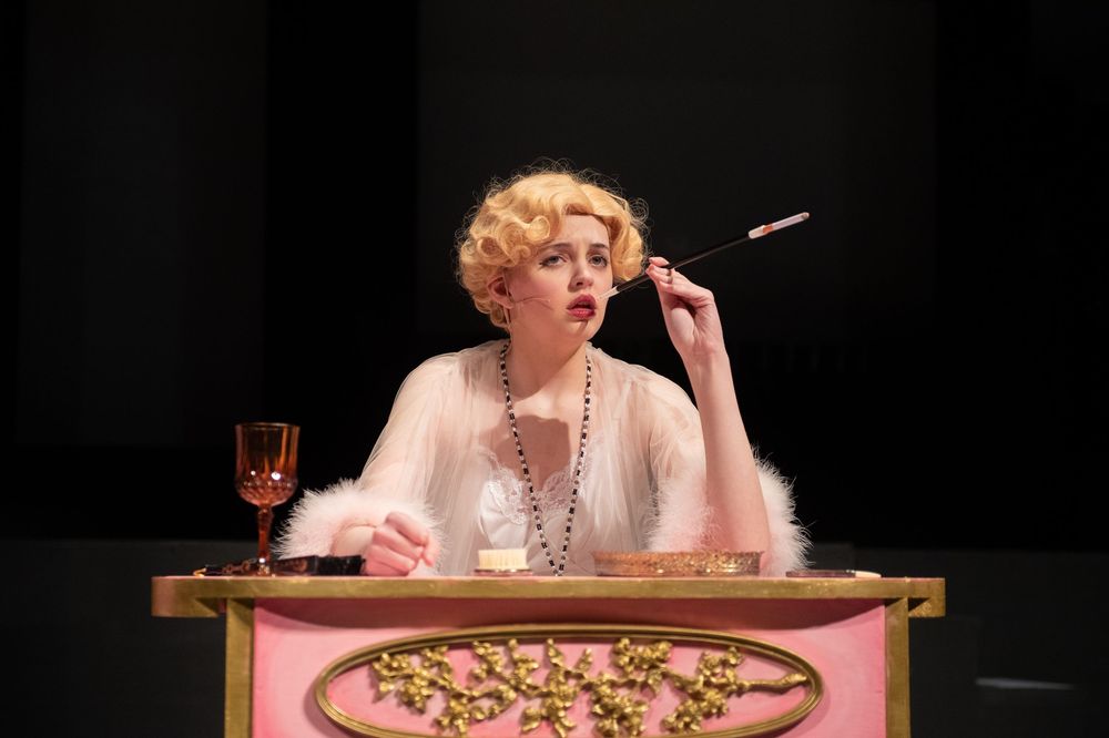 A performer in a white dress and pearls sits at a pink table while speaking into a microphone during a theatrical performance.