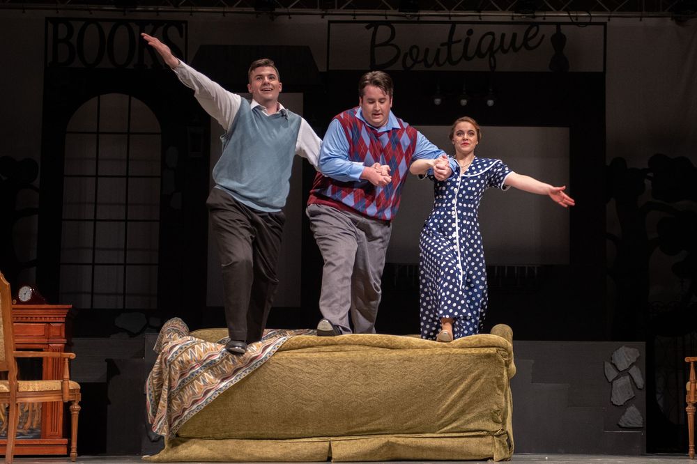 Three performers stand triumphantly on a couch during what appears to be a theatrical stage production.