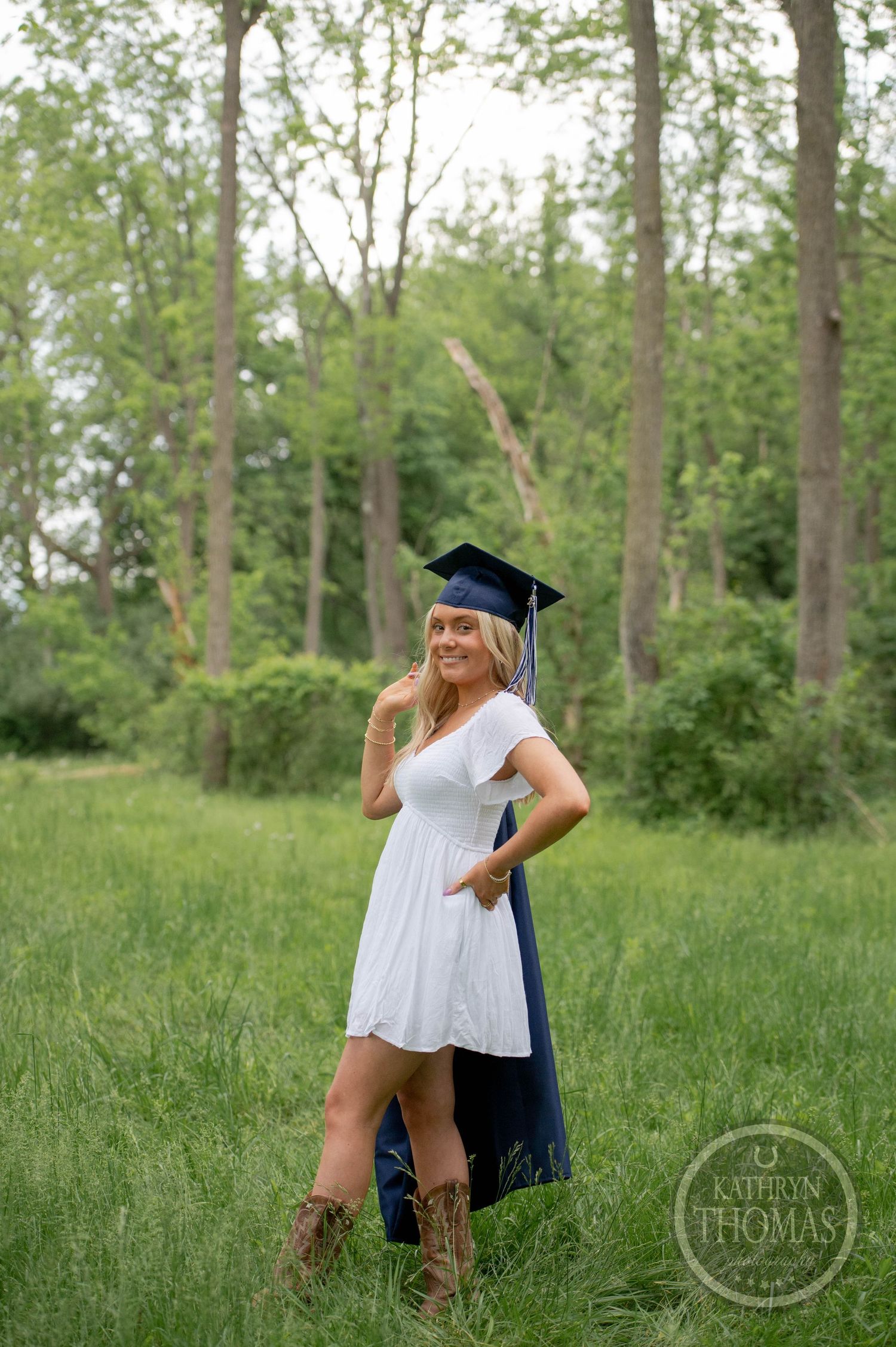 A graduate wearing a white summer dress, cowboy boots and graduation cap poses in a wooded outdoor setting.