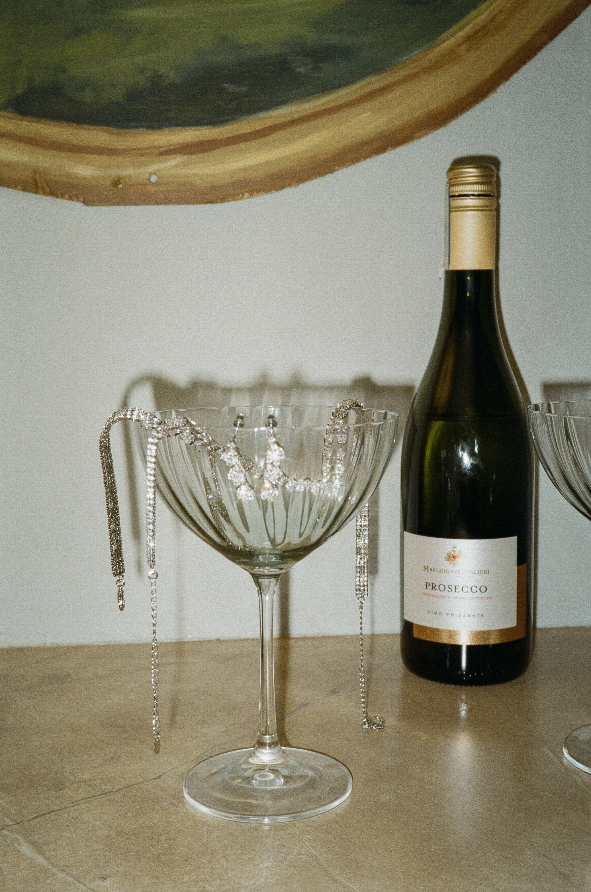 A bottle of Prosecco stands next to an elegant crystal coupe glass adorned with hanging crystal beads on a beige surface.