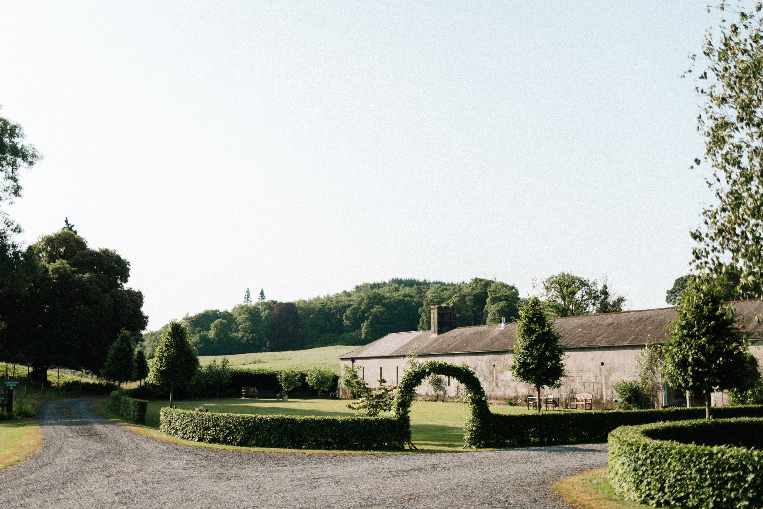 A stone archway leads to a rural country estate with manicured hedges and rolling hills in the background.
