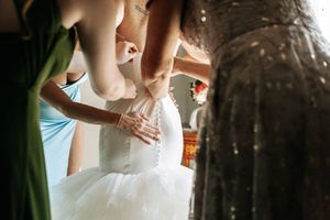 A wedding scene shows hands helping to adjust a white tulle bridal gown in soft natural lighting.