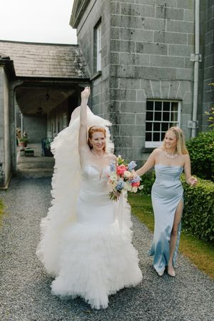 A bride in a flowing white wedding dress walks on a gravel path with a bridesmaid in a light blue gown beside a stone building.