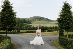 A scenic country road framed by evergreen trees leads to rolling green hills with a figure in white standing in the middle path.