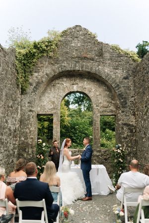 Intimate wedding ceremony takes place under a rustic stone archway adorned with greenery and white flowers at historic ruins.