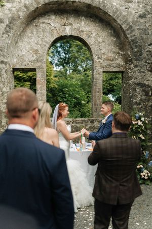 A wedding ceremony takes place under a stone archway surrounded by lush greenery at a rustic outdoor venue.