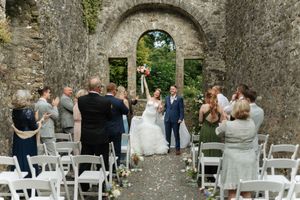 Wedding ceremony taking place in stone ruins with white chairs and castle archway framing the scenic backdrop.