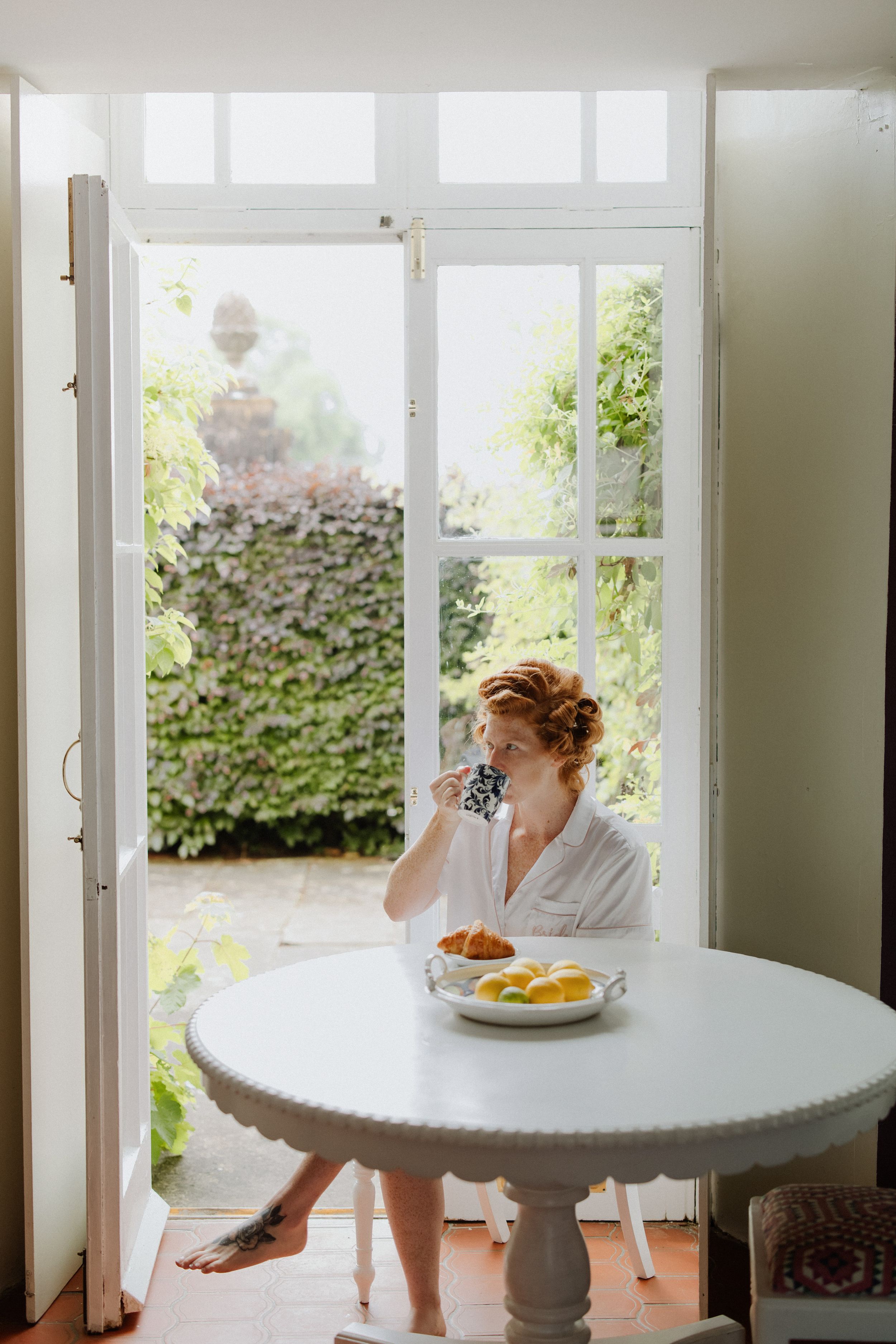 Soft morning light streams through French doors onto a white round table where someone enjoys breakfast in a robe.