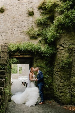 A bride and groom share an intimate moment in a stone archway covered with lush green vines and foliage.