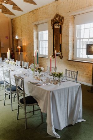 Elegant dining table set with white linens, silver chairs and candles in a rustic brick-walled room with large windows.