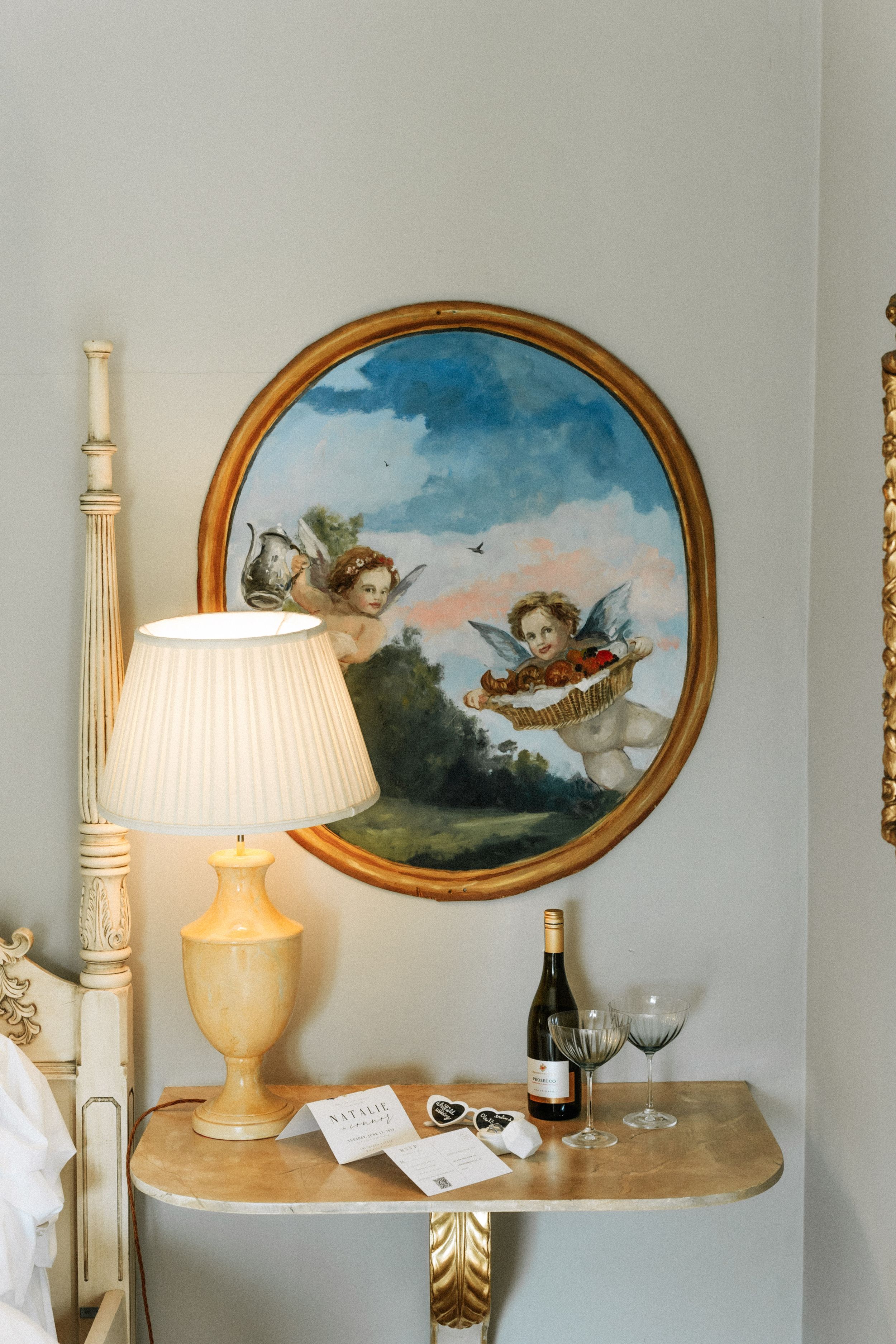 Elegant bedside table with antique lamp and round framed cherub artwork, beside wine and glasses on gold accented table.