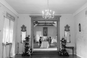 Black and white interior view of an elegant wedding ceremony through a doorway with a crystal chandelier and decorative trees.