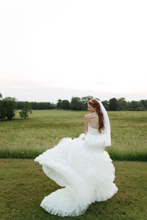 A bride in a flowing white wedding gown twirls in a scenic green field during a romantic outdoor photo session.