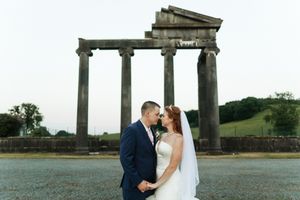 Newlyweds celebrate their wedding at an ancient Roman ruins site with stone pillars against a green hillside background.