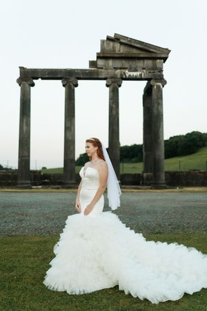 A bride in a flowing white gown with long train poses in front of ancient Roman stone columns at an outdoor location.