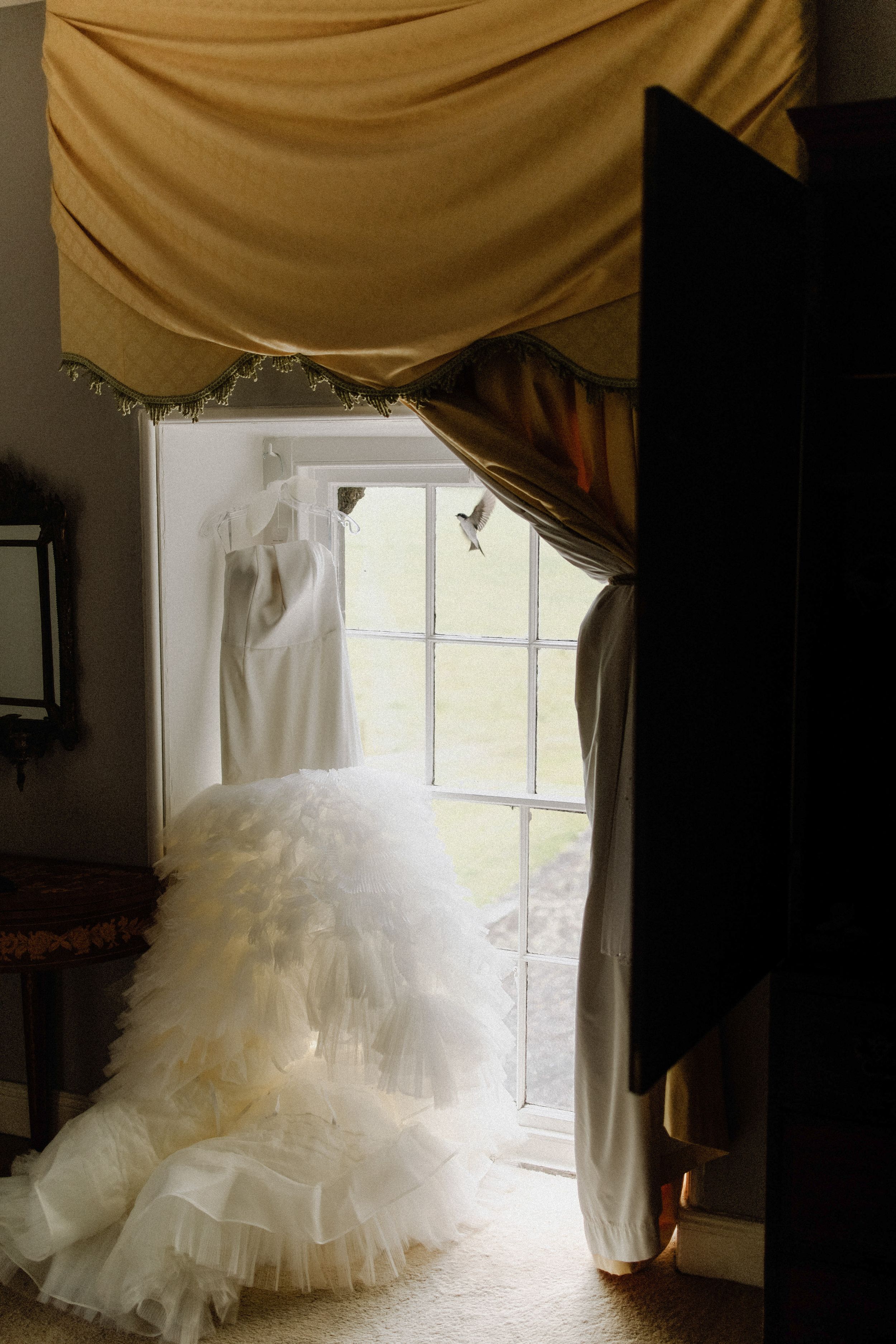 A flowing white wedding dress with ruffled tulle skirt hangs by a sunlit window beneath gold draped fabric.