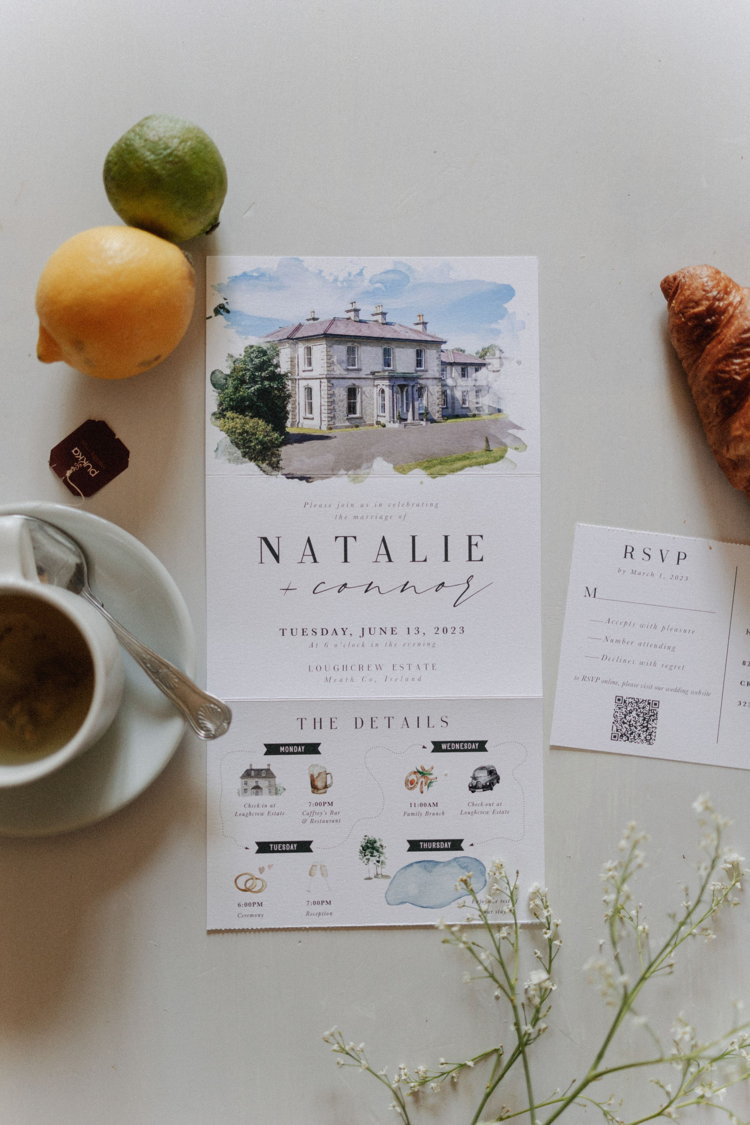 Elegant wedding invitation featuring watercolor venue illustration displayed with citrus fruits, coffee, and delicate white flowers.