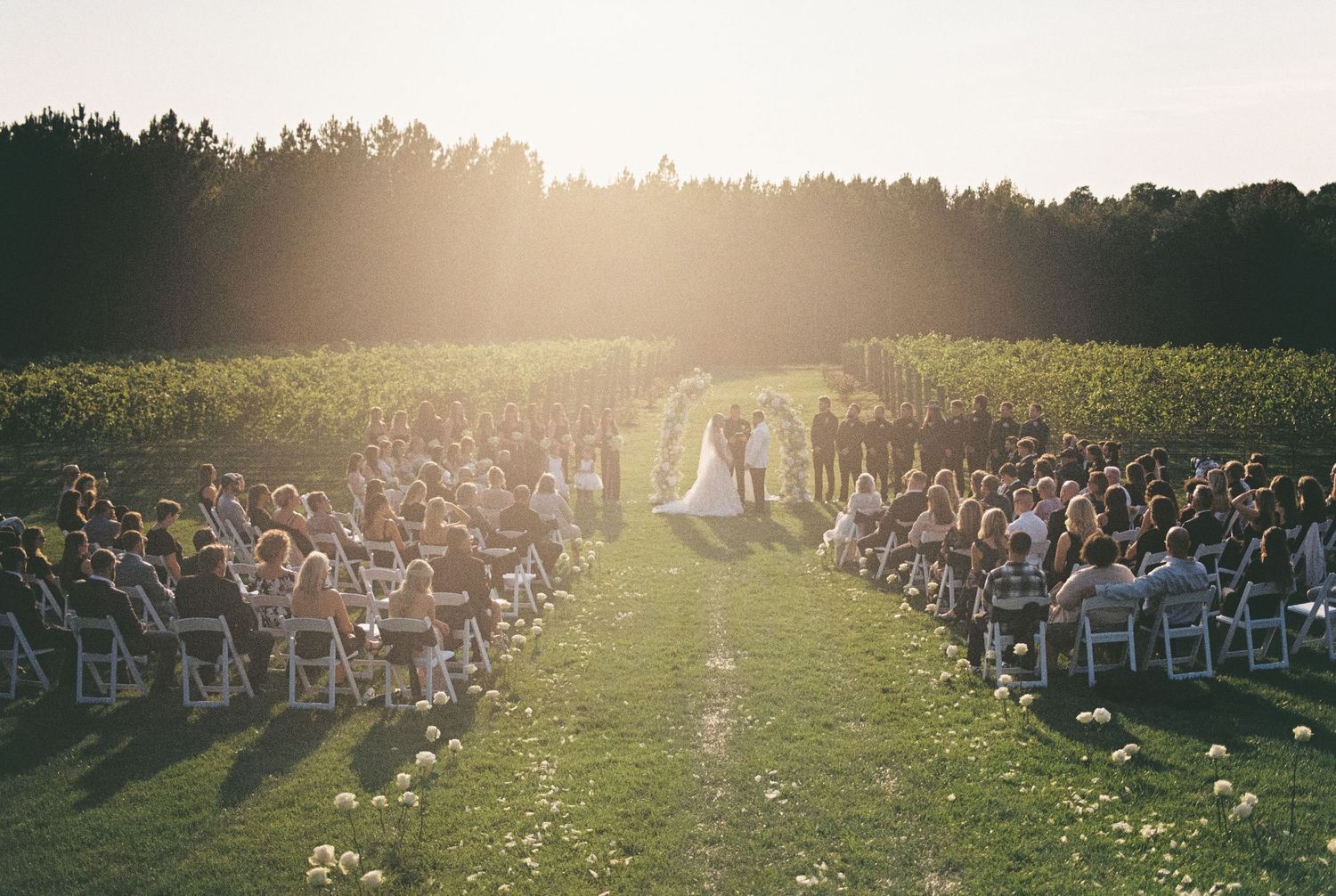 Outdoor wedding ceremony in sunlit field with rows of white chairs and guests gathering at golden hour.