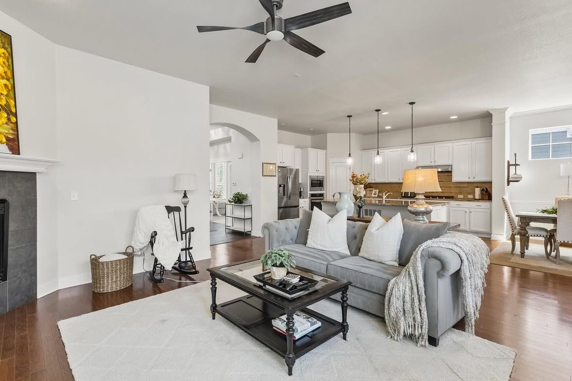 Open concept living room with gray sofa, coffee table and modern decor flowing into bright kitchen with ceiling fan.
