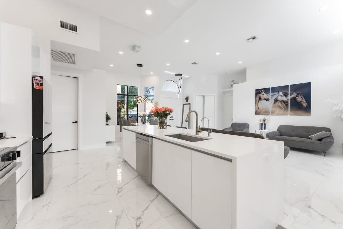 Modern white kitchen with sleek island, marble floors, stainless appliances and grey sofa in open floor plan design.
