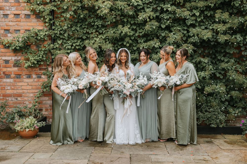 Bridal party in sage green dresses poses outdoors along brick wall for wedding photos.