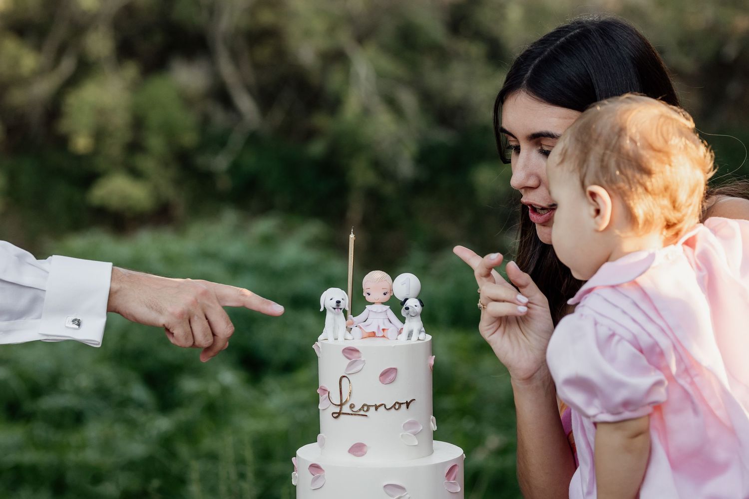 Someone blows out candles on a birthday cake with white frosting and floral decorations while another person points at the cake.