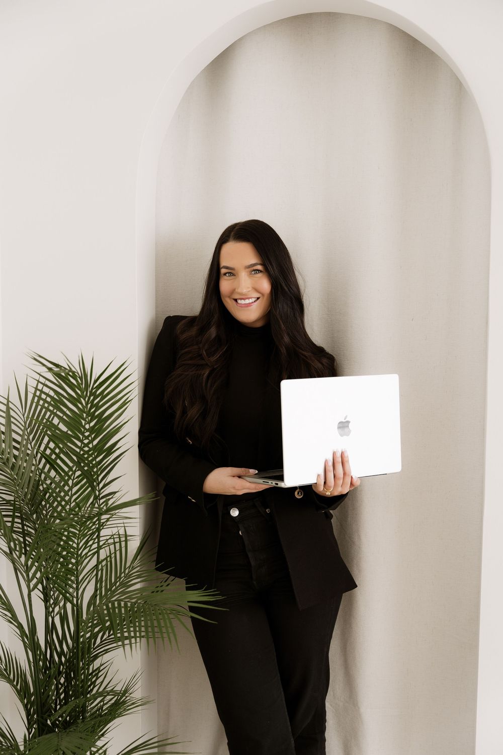 Professional in black attire stands in modern white archway with palm plant and laptop.