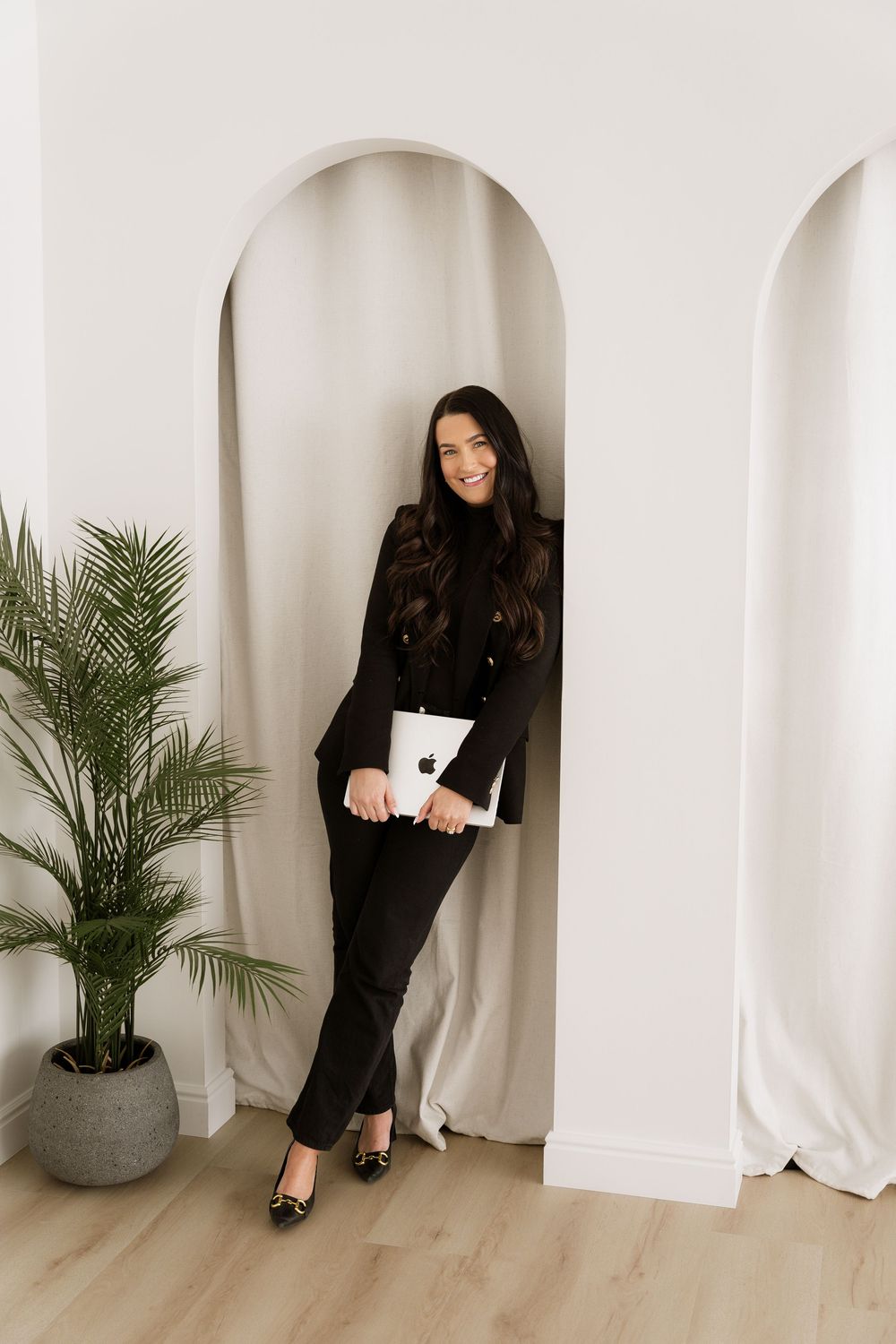 Professional holding laptop while standing in arched doorway with palm plant decor.